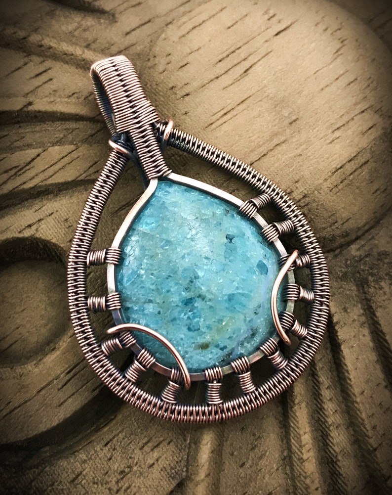 K&ouml;nnte beinhalten: Ein mit Kupferdraht umwickelter Anh&auml;nger mit einem hellblauen Edelstein. Der Anh&auml;nger hat die Form einer Tr&auml;ne und ein filigranes Drahtdesign.