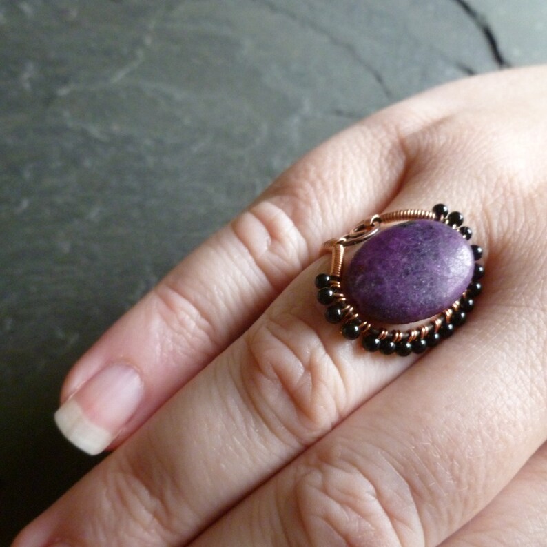Op de afbeelding: Een ring met een paarse edelsteen, zwarte kralen en een wikkeling van koperdraad. De ring zit aan een vinger.