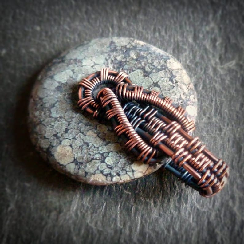 Puede incluir: Un colgante de alambre de cobre y negro con un dise&ntilde;o tejido. El colgante est&aacute; sobre una piedra gris y blanca.