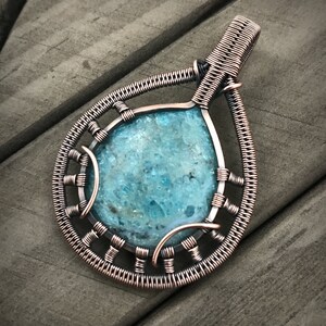 K&ouml;nnte beinhalten: Ein mit Kupferdraht umwickelter Anh&auml;nger mit einem blaugr&uuml;nen Edelstein. Der Anh&auml;nger hat die Form einer Tr&auml;ne und ein filigranes, kompliziertes Design.