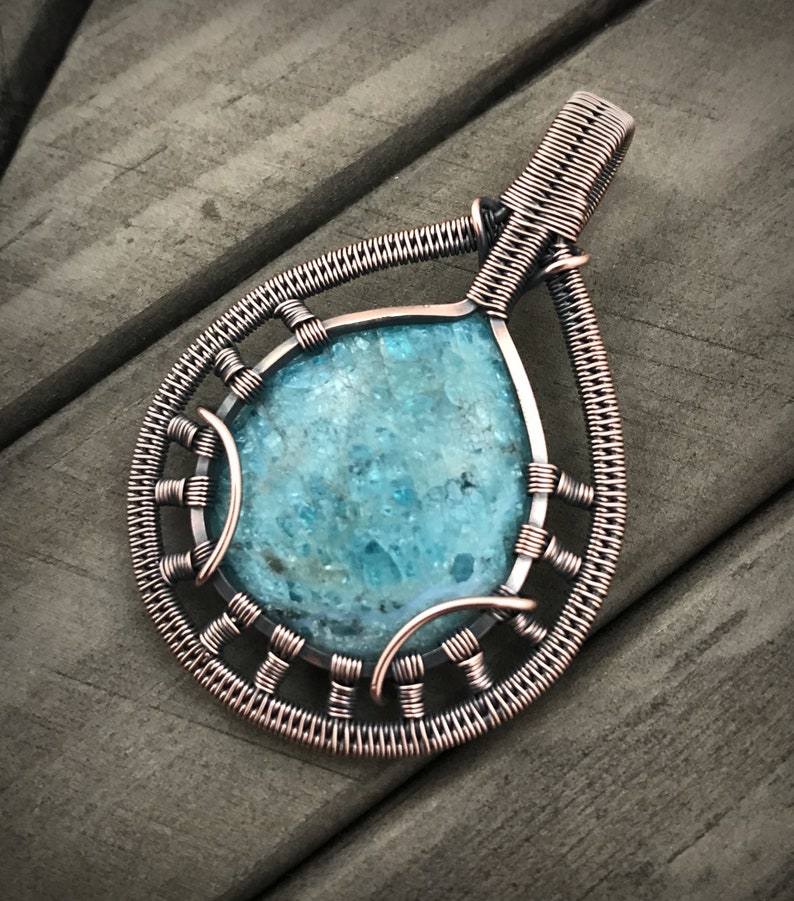 K&ouml;nnte beinhalten: Ein mit Kupferdraht umwickelter Anh&auml;nger mit einem blaugr&uuml;nen Edelstein. Der Anh&auml;nger hat die Form einer Tr&auml;ne und ein filigranes, kompliziertes Design.