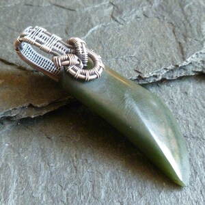 Puede incluir: Un colgante de jade verde con una fianza envuelta en alambre de plata. El colgante tiene la forma de una l&aacute;grima larga y delgada.