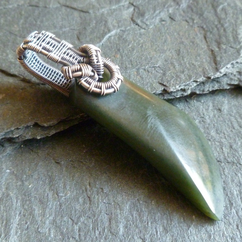 Puede incluir: Un colgante de jade verde con una fianza envuelta en alambre de plata. El colgante tiene la forma de una l&aacute;grima larga y delgada.