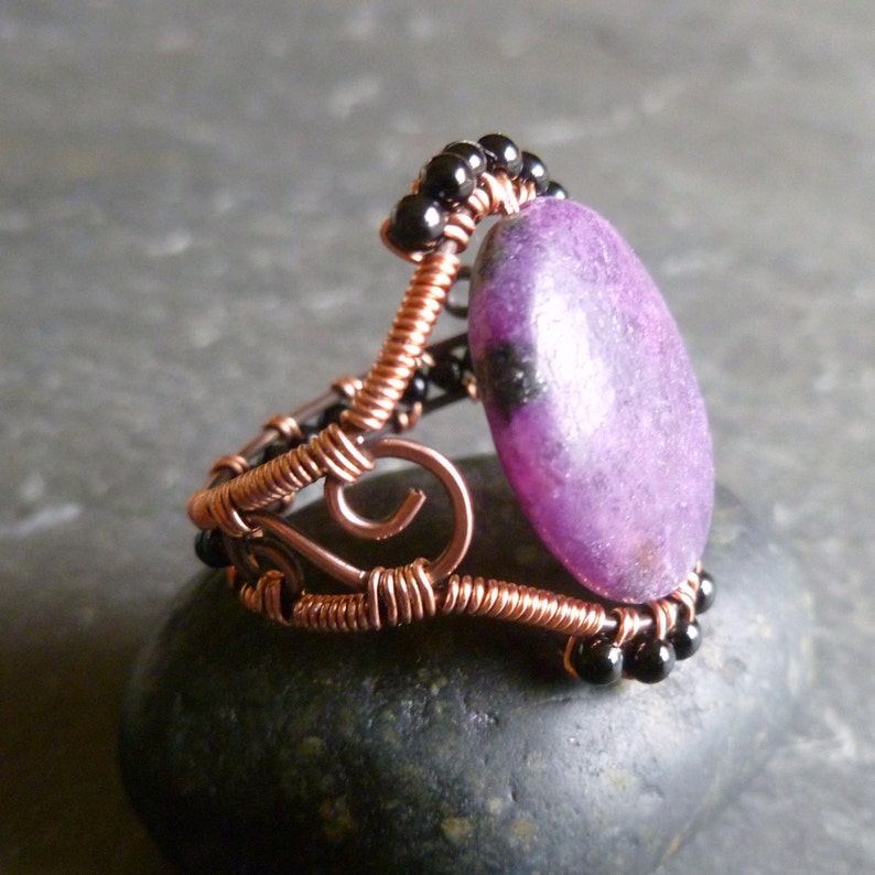 Op de afbeelding: Een ring met een paarse edelsteen, koperdraad en zwarte kralen. De ring is handgemaakt en heeft een uniek ontwerp.