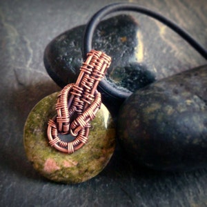 Puede incluir: Un colgante de alambre de cobre envuelto con una piedra verde y rosa. El colgante est&aacute; colgado de un cord&oacute;n negro.