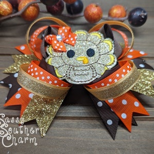 May include: A brown, orange, and gold glitter hair bow with a sparkly silver turkey applique. The bow has white polka dots on the orange and brown ribbons. The turkey has a yellow beak and black eyes. The bow is tied with a red ribbon with a rhinestone in the center. The text "Sassy Southern Charm" is visible in the bottom left corner of the image.
