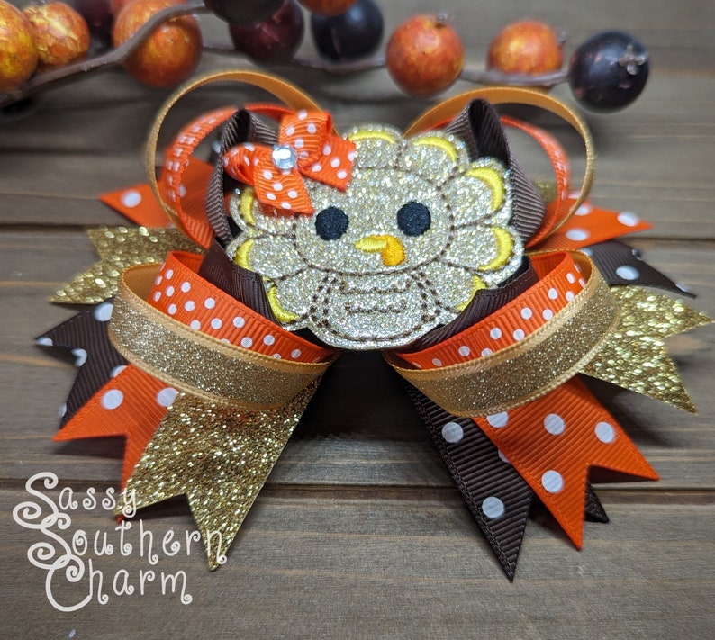 May include: A brown, orange, and gold glitter hair bow with a sparkly silver turkey applique. The bow has white polka dots on the orange and brown ribbons. The turkey has a yellow beak and black eyes. The bow is tied with a red ribbon with a rhinestone in the center. The text "Sassy Southern Charm" is visible in the bottom left corner of the image.