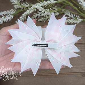 Puede incluir: Un lazo grande para el cabello blanco con detalles en rosa. El lazo presenta un dise&ntilde;o en capas con bordes puntiagudos y un clip met&aacute;lico en la parte posterior. El texto "Sassy Southern Charm" es visible en la parte inferior izquierda.