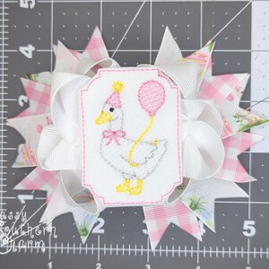 Puede incluir: Un lazo para el pelo hecho a mano con un dise&ntilde;o blanco y rosa. El lazo presenta un parche bordado de un ganso sosteniendo un globo y usando un gorro de fiesta. El texto "Sassy Southern Charm" es visible.