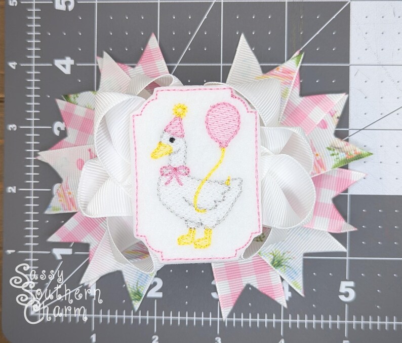 Puede incluir: Un lazo para el pelo hecho a mano con un dise&ntilde;o blanco y rosa. El lazo presenta un parche bordado de un ganso sosteniendo un globo y usando un gorro de fiesta. El texto "Sassy Southern Charm" es visible.