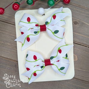 May include: Two white Christmas light hair bows with red and green accents. The bows are on a white card with the text "Sassy Southern Charm".