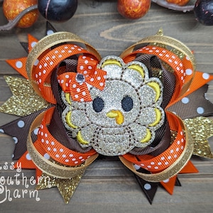 May include: A brown, orange, and gold glitter hair bow with a sparkly silver turkey applique. The turkey has a yellow beak and black eyes. The bow is decorated with white polka dots.