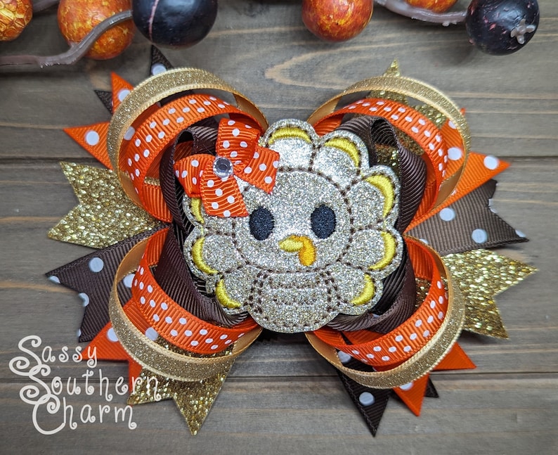 May include: A brown, orange, and gold glitter hair bow with a sparkly silver turkey applique. The turkey has a yellow beak and black eyes. The bow is decorated with white polka dots.
