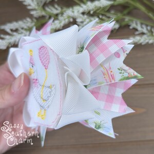 Puede incluir: Un lazo para el cabello hecho a mano con una base blanca y un ganso bordado sosteniendo un globo rosa. El lazo tiene capas de cintas blancas, rosas a cuadros y florales. El texto "Sassy Southern Charm" es visible.