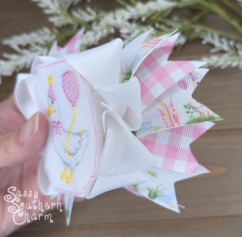 Puede incluir: Un lazo para el cabello hecho a mano con una base blanca y un ganso bordado sosteniendo un globo rosa. El lazo tiene capas de cintas blancas, rosas a cuadros y florales. El texto "Sassy Southern Charm" es visible.