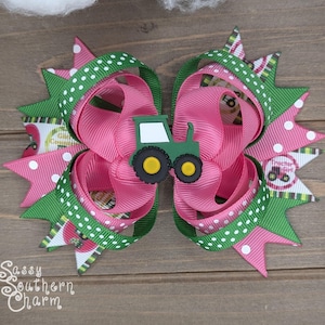 Puede incluir: Un lazo para el pelo rosa y verde con un amuleto de tractor verde. El lazo est&aacute; hecho de cinta con lunares blancos y un patr&oacute;n de rayas verdes y blancas. El amuleto de tractor tiene un parabrisas blanco y ruedas amarillas.