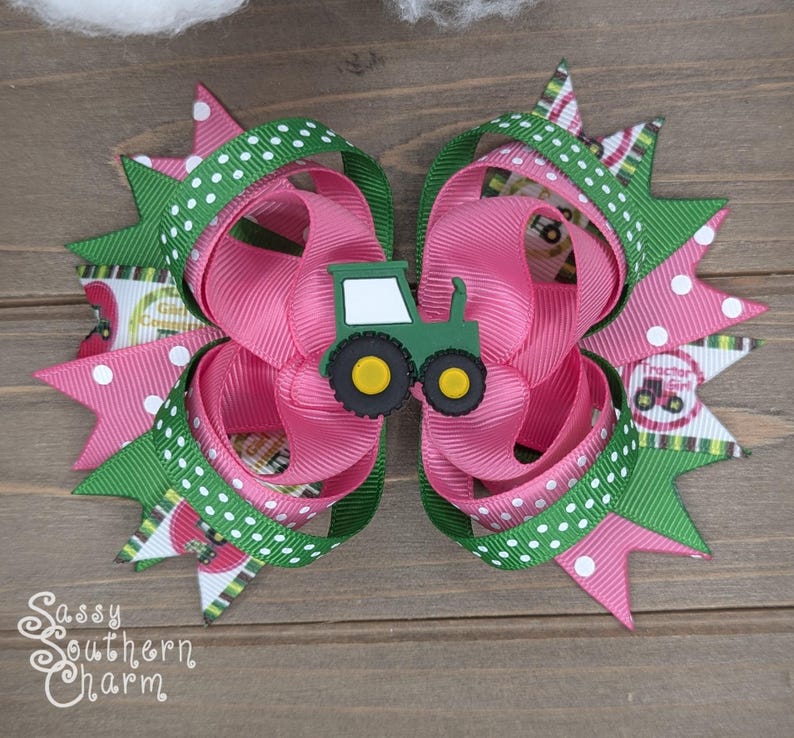 Puede incluir: Un lazo para el pelo rosa y verde con un amuleto de tractor verde. El lazo est&aacute; hecho de cinta con lunares blancos y un patr&oacute;n de rayas verdes y blancas. El amuleto de tractor tiene un parabrisas blanco y ruedas amarillas.