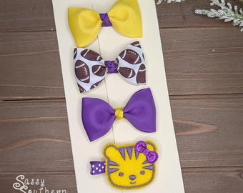 May include: A set of four hair accessories on a display card. The set includes a solid yellow bow with a purple center, a white bow with a brown football pattern, a solid purple bow with a yellow center, and a yellow tiger face felt clip.