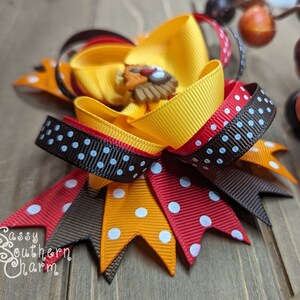 K&ouml;nnte beinhalten: Eine handgefertigte Haarschleife mit gelben, orangefarbenen, roten und braunen B&auml;ndern. Die Schleife hat eine Truthahn-f&ouml;rmige Verzierung in der Mitte und wei&szlig;e Tupfen. Der Text "Sassy Southern Charm" ist sichtbar.