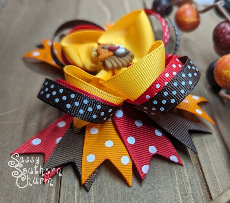 K&ouml;nnte beinhalten: Eine handgefertigte Haarschleife mit gelben, orangefarbenen, roten und braunen B&auml;ndern. Die Schleife hat eine Truthahn-f&ouml;rmige Verzierung in der Mitte und wei&szlig;e Tupfen. Der Text "Sassy Southern Charm" ist sichtbar.