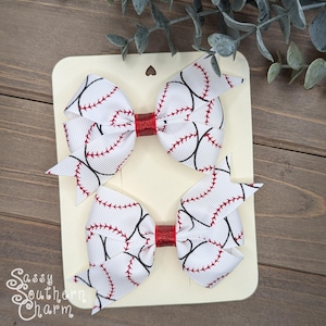 May include: Two white hair bows with a baseball print and red glitter centers. The bows are on a white card with the text "Sassy Southern Charm".