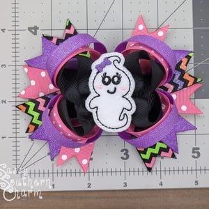 Peut inclure: Un n&oelig;ud pour cheveux violet, rose, noir et vert avec une applique en feutre blanc en forme de fant&ocirc;me. Le fant&ocirc;me a un visage souriant avec deux yeux noirs et un n&oelig;ud violet sur la t&ecirc;te.