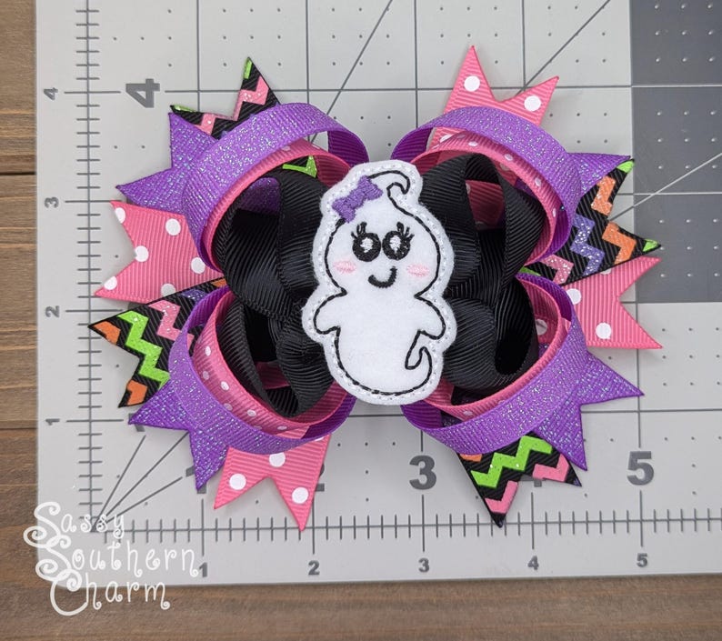 Peut inclure: Un n&oelig;ud pour cheveux violet, rose, noir et vert avec une applique en feutre blanc en forme de fant&ocirc;me. Le fant&ocirc;me a un visage souriant avec deux yeux noirs et un n&oelig;ud violet sur la t&ecirc;te.