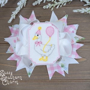Puede incluir: Un lazo decorativo para el cabello con un estampado de cuadros blancos y rosas y un bordado de un ganso sosteniendo un globo. El ganso lleva un gorro de fiesta. El lazo tiene varias capas y el texto "Sassy Southern Charm".