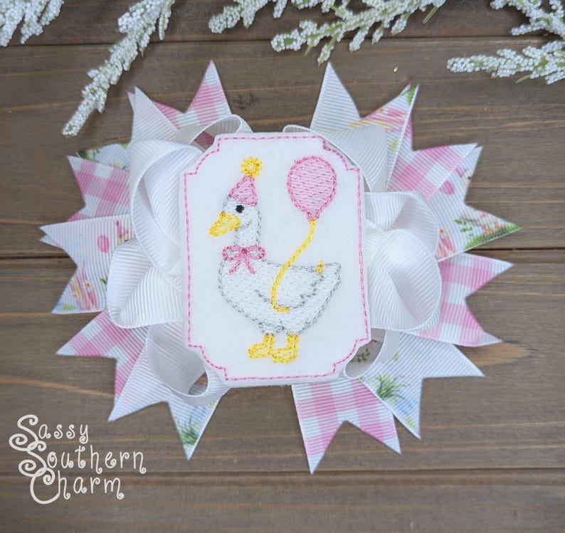 Puede incluir: Un lazo decorativo para el cabello con un estampado de cuadros blancos y rosas y un bordado de un ganso sosteniendo un globo. El ganso lleva un gorro de fiesta. El lazo tiene varias capas y el texto "Sassy Southern Charm".
