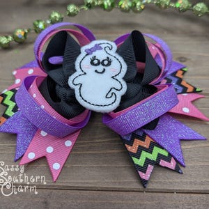 Peut inclure: Un n&oelig;ud pour cheveux rose, violet, noir et vert avec une applique de fant&ocirc;me blanc. Le fant&ocirc;me a un visage souriant et un n&oelig;ud violet sur la t&ecirc;te. Le n&oelig;ud est compos&eacute; de plusieurs couches de ruban et pr&eacute;sente un motif en chevron.