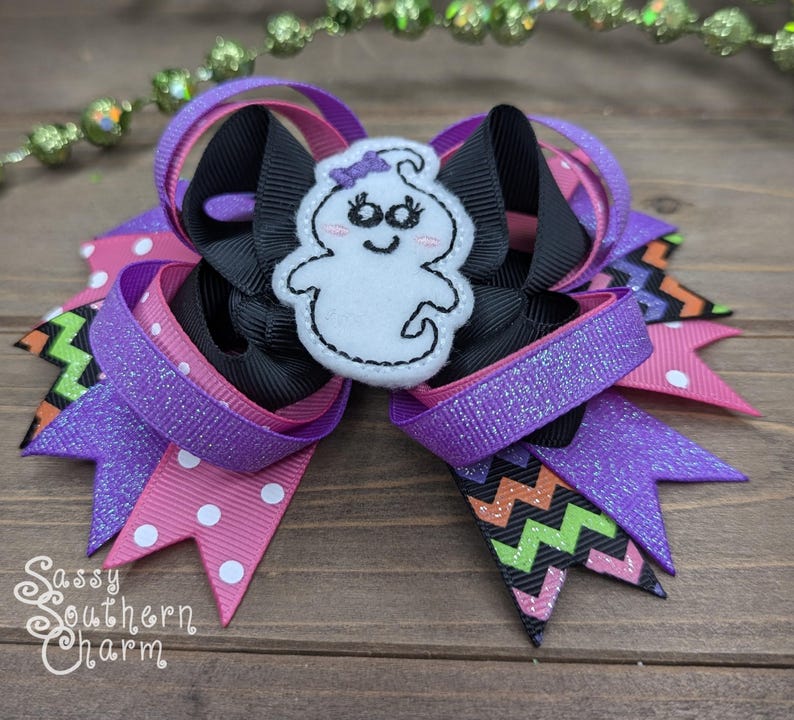 Peut inclure: Un n&oelig;ud pour cheveux rose, violet, noir et vert avec une applique de fant&ocirc;me blanc. Le fant&ocirc;me a un visage souriant et un n&oelig;ud violet sur la t&ecirc;te. Le n&oelig;ud est compos&eacute; de plusieurs couches de ruban et pr&eacute;sente un motif en chevron.