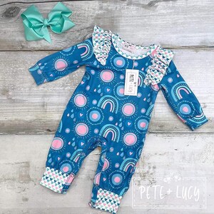 Puede incluir: Un body azul para bebé con un estampado de soles rosas, arcoíris y gotas de lluvia. El body tiene mangas largas, un cuello con volantes y puños con un estampado de corazones. Un lazo turquesa a juego está en el fondo. La marca "PETE + LUCY" es visible.