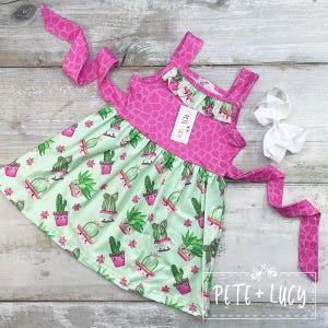 Puede incluir: Un vestido sin mangas con un corpiño y tirantes con estampado de jirafa rosa, y una falda verde menta estampada con cactus de dibujos animados en macetas rosas. También hay un lazo blanco y una corbata rosa. El vestido tiene la marca "PETE + LUCY".