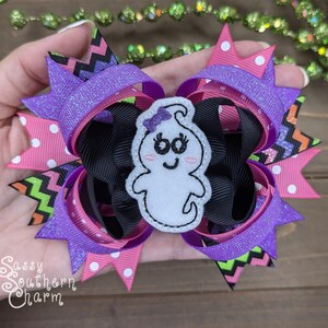 Peut inclure: Un n&oelig;ud pour cheveux violet, rose, noir et vert avec une applique de fant&ocirc;me blanc. Le fant&ocirc;me a un sourire et des yeux noirs et un n&oelig;ud rose sur la t&ecirc;te.