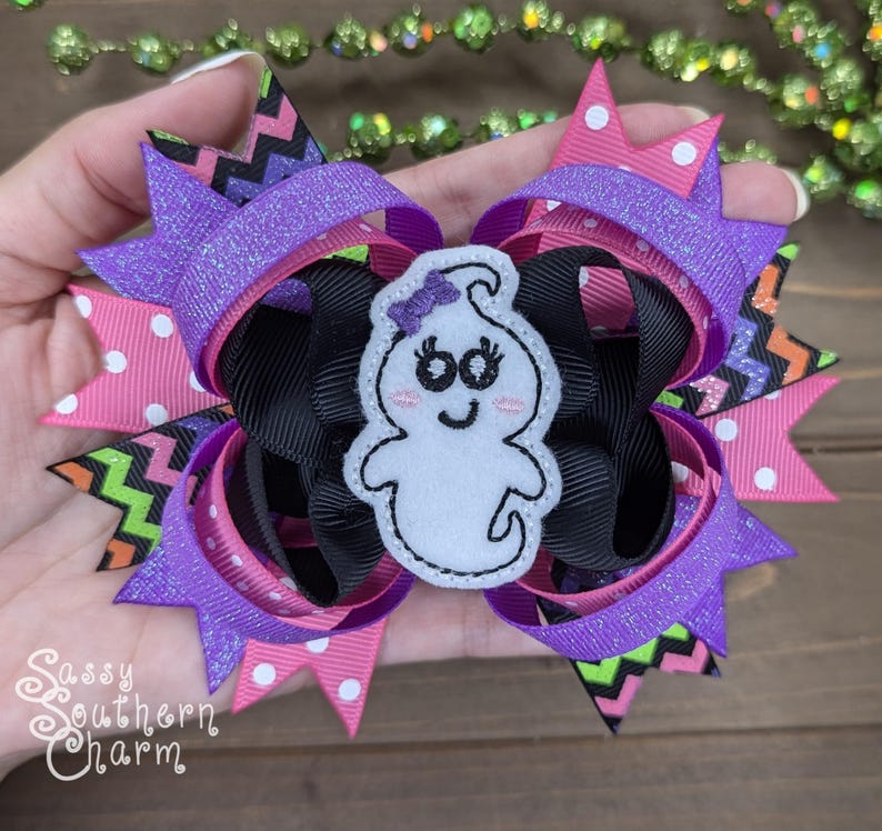 Peut inclure: Un n&oelig;ud pour cheveux violet, rose, noir et vert avec une applique de fant&ocirc;me blanc. Le fant&ocirc;me a un sourire et des yeux noirs et un n&oelig;ud rose sur la t&ecirc;te.
