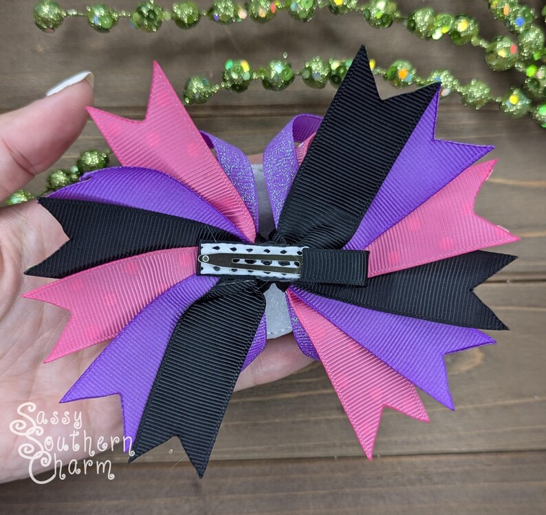 Peut inclure: Un n&oelig;ud pour cheveux violet, rose et noir avec une pince &agrave; l'arri&egrave;re. Le n&oelig;ud est fait de ruban et a un centre scintillant.