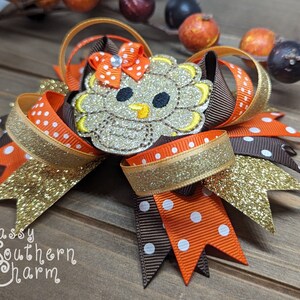 May include: A brown, orange, and gold glitter hair bow with a felt turkey applique. The turkey has a brown body, orange beak, and white eyes. The bow is decorated with white polka dots.