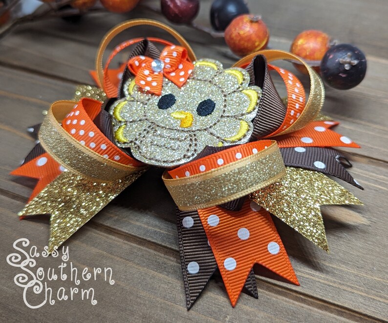 May include: A brown, orange, and gold glitter hair bow with a felt turkey applique. The turkey has a brown body, orange beak, and white eyes. The bow is decorated with white polka dots.