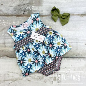 Puede incluir: Un body de bebé sin mangas con un estampado floral de margaritas en azul, blanco, rosa y negro. El body tiene una cintura y una parte inferior a rayas. Un lazo verde oliva se coloca junto al body. La etiqueta dice "PETE + LUCY".