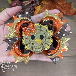 May include: A brown, orange, and gold glitter hair bow with a glittery brown turkey applique in the center. The turkey has a yellow beak and white eyes.