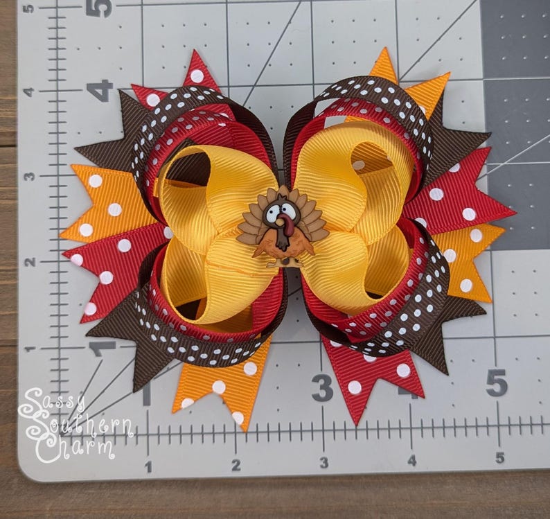 K&ouml;nnte beinhalten: Eine festliche Haarschleife mit einem Truthahn-Emblem. Die Schleife besteht aus geschichteten B&auml;ndern in Gelb, Rot, Braun und Orange mit wei&szlig;en Punkten. Die Schleife ist etwa 13 cm breit, ideal f&uuml;r Thanksgiving oder herbstliche Anl&auml;sse.