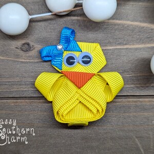 May include: A yellow chick hair bow with blue and white accents. The chick has large black eyes and an orange beak. The bow is made of ribbon and is attached to a clip.