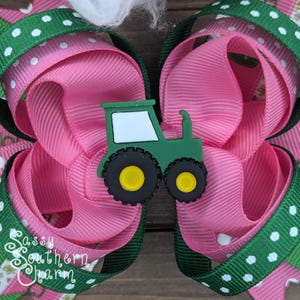 Puede incluir: Un lazo para el pelo rosa y verde con un amuleto de tractor verde. El tractor es de color verde oscuro con detalles blancos y ruedas amarillas. El lazo est&aacute; hecho de dos capas de cinta rosa con lunares blancos y cinta verde con lunares blancos.