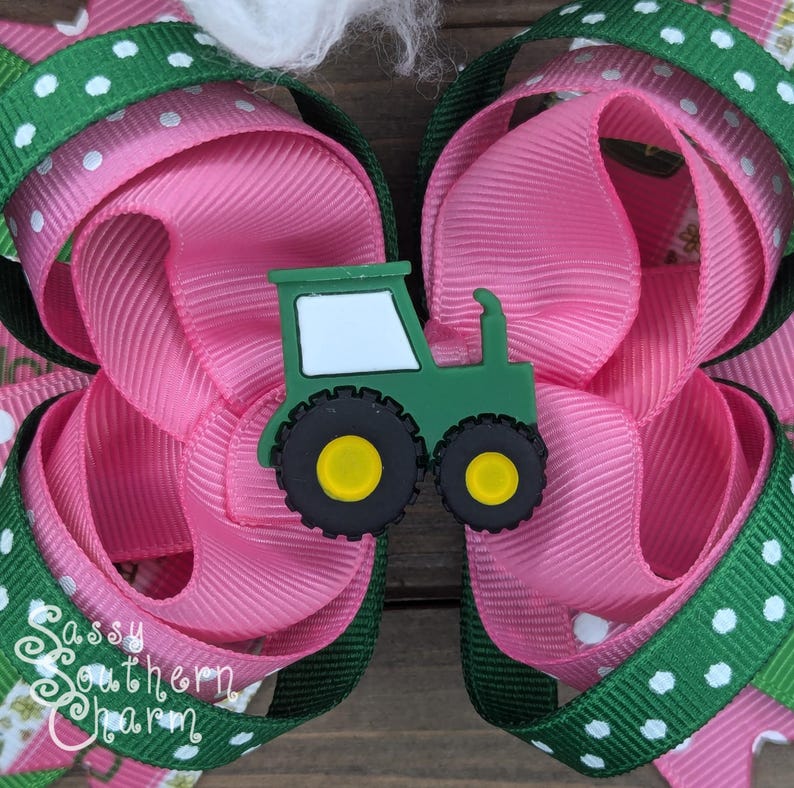 Puede incluir: Un lazo para el pelo rosa y verde con un amuleto de tractor verde. El tractor es de color verde oscuro con detalles blancos y ruedas amarillas. El lazo est&aacute; hecho de dos capas de cinta rosa con lunares blancos y cinta verde con lunares blancos.