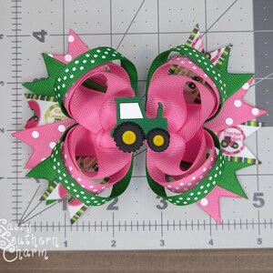 Puede incluir: Un lazo para el pelo rosa y verde con un amuleto de tractor verde. El lazo est&aacute; hecho de cinta con lunares blancos y un patr&oacute;n de cuadros verdes y blancos. El amuleto de tractor tiene una cabina blanca y ruedas amarillas.