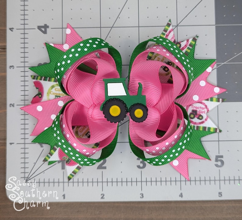 Puede incluir: Un lazo para el pelo rosa y verde con un amuleto de tractor verde. El lazo est&aacute; hecho de cinta con lunares blancos y un patr&oacute;n de cuadros verdes y blancos. El amuleto de tractor tiene una cabina blanca y ruedas amarillas.