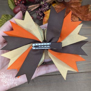 May include: A brown, gold, and orange hair bow with a black and white polka dot alligator clip. The bow is made of grosgrain ribbon and has a layered, ruffled design.