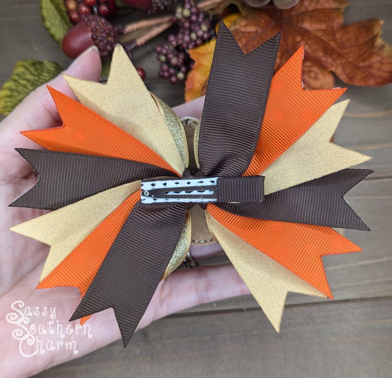 May include: A brown, gold, and orange hair bow with a black and white polka dot alligator clip. The bow is made of grosgrain ribbon and has a layered, ruffled design.