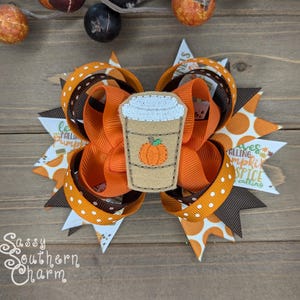 Puede incluir: Un lazo para el pelo marrón y naranja con lunares blancos y una aplicación de fieltro de una taza de café con una calabaza dentro. El lazo tiene el texto "Leaves falling pumpkin spice calling".