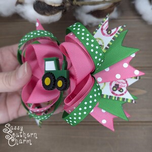 Puede incluir: Un lazo para el pelo rosa y verde con lunares blancos y un amuleto de tractor verde. El lazo est&aacute; hecho de cinta y est&aacute; atado en un nudo grande y esponjoso.